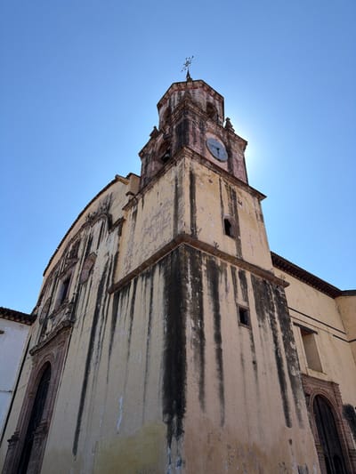 The image captures a low-angle perspective of an old, grand architectural structure, likely a church or a historical building, set against a vast, clear blue sky. The building appears to lean significantly towards the right, giving the composition a dynamic and somewhat tilted feel, suggesting the photo was taken while looking sharply upwards.
The central feature is a prominent square tower, possibly a bell tower, topped with a small, ornate cross or weather vane. Below this, a circular clock face is visible on one side of the tower, though the exact time is not clearly discernible. The tower and the adjacent main body of the building are constructed from a light-colored material, possibly stucco or stone, which has aged considerably. Dark, irregular streaks and patches are prevalent across the surfaces, indicating significant weathering, dirt, or water damage accumulated over time. These dark stains run both horizontally and vertically, creating a textured and somewhat weathered appearance.
The main building extends to the right of the tower, showcasing more of the light-colored walls and dark streaking. Portions of a terracotta-colored tiled roof are visible along the top edges of both the tower and the main structure. Several small, dark, rectangular or arched openings, possibly windows or vents, are scattered across the building's facade. On the far right, a larger arched window with dark bars or a grille can be seen, hinting at more elaborate architectural details further along the building.
The sky is a vivid and uniform clear blue, occupying the upper and left portions of the frame. The sun is positioned directly behind the tower, creating a bright halo or lens flare effect that slightly obscures the very top edge of the tower but intensely illuminates the scene, highlighting the textures and colors of the building. This strong backlighting suggests a sunny day, likely mid-morning or afternoon. There are no people, vehicles, or discernible text visible in the image. The overall impression is one of ancient architecture standing majestically under a bright, open sky.
