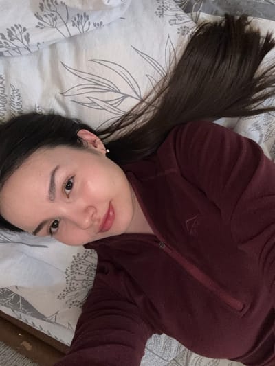 An individual, identified as female, is observed in an indoor environment, specifically a bed, located in the city of Bayronovka, Russia. She lies in a supine position, angled with her head towards the upper left of the frame. Her long, dark hair is spread across a light-colored sheet featuring a repeating grey botanical pattern. The individual possesses fair skin, dark eyes, and full lips, displaying a subtle, direct gaze at the camera. She is attired in a maroon fleece top, which includes a partial zipper closure and a light-colored, stylized 'mountain' or 'arrow' logo embroidered on the left chest. The left ear is visible, adorned with a small hoop earring and a stud earring. A wooden element is partially visible at the bottom left of the image.