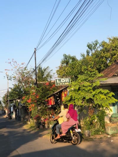 A motorcycle with two occupants is traveling down a paved road in Kalidawir, Indonesia. The driver is wearing a yellow shirt and blue jeans, while the passenger is wearing a pink hijab and a pink dress. The motorcycle is black and blue. On the right side of the road, there is a building with a tiled roof and a green door. There are also trees and bushes with red and orange flowers. A sign with the text "LDII" and an arrow pointing to the right is visible on the left side of the road, above a small building. The sky is clear and blue, and the sun is casting shadows on the road, indicating it is either morning or late afternoon. Electrical wires are strung across the sky, supported by utility poles.