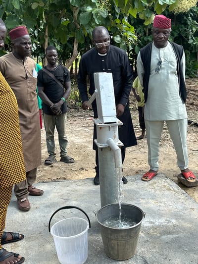 In Afogo, Nigeria, a group of men are gathered around a manual water pump. The pump, a silver metal cylinder with a lever arm and a spout, is actively dispensing water into a metal bucket. The ground is dusty earth, with patches of concrete where the pump is situated. Several men are dressed in traditional attire, including a man in a brown tunic and a red cap, and another in a light blue tunic and a dark vest, also wearing a red cap. A man in a black shirt and glasses appears to be operating the pump, while others observe. A clear plastic bucket also sits nearby. The background features lush green foliage, suggesting an outdoor, possibly rural or semi-rural, setting.