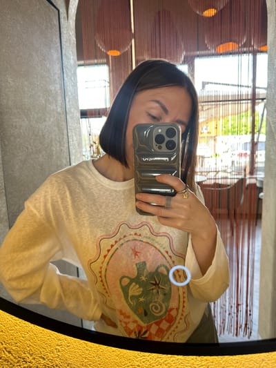 A female subject with dark brown bob-cut hair is depicted taking a mirror selfie in an indoor setting in Усады, Russia. Her reflection shows her holding an iPhone with a ribbed, translucent grey case displaying "VVV" and other text. She wears a light-colored, long-sleeved t-shirt featuring a circular graphic with a stylized pitcher, stars, and checkered patterns, and partially visible text "Il Cielo Picasso Il". Multiple rings are on her right hand: a multi-band silver-toned ring and a gold-toned ring with a small flower charm. The background, visible through the mirror, is a blurred interior space, likely a restaurant, characterized by vertical reddish-brown decorative strands, spherical orange light fixtures, and large windows revealing a bright outdoor scene with green foliage. The bottom edge of the mirror shows a textured gold-yellow surface.