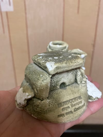 The image shows a close-up of a person's hand holding a small, weathered, light-colored ceramic figurine of a frog. The figurine has a slightly mottled, greenish-brown patina with white paint chips peeling off in several places, suggesting age or wear. The frog's features are somewhat stylized and rounded.
Visible text on the base of the figurine, oriented in the Russian alphabet, appears to be "ЧЕЛЯБИНСК" (Chelyabinsk), followed by "Мастер Еломан" (Master Yeloman). Below this, there's potentially more text that is too blurry to decipher accurately.
The background is a light brown, textured surface, possibly wood paneling, with faint horizontal lines. The lighting is diffuse, and no specific time of day or weather conditions can be determined. The scene is a close-up, focusing entirely on the figurine and the hand holding it. The context provided indicates the location as Novaya Usman', Russia, which is external information not directly visible in the image itself.