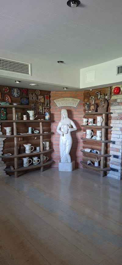 This is a full-shot, eye-level indoor photograph of the Katharine Pottery (Käsitöölise Puhak Katharine) in Angla, Estonia. The room has a rustic interior design with exposed brick walls and wooden shelves displaying pottery. In the center of the image, there is a large, white marble statue of a woman holding a bowl with a starburst pattern. The statue stands on a rectangular pedestal.
To the left of the statue, there are several wooden shelves filled with various ceramic items, including pitchers, bowls, and decorative plates. Some of these items are adorned with floral motifs.
To the right of the statue, more wooden shelves are visible, also displaying pottery, including mugs, pitchers, and decorative objects. There is a red fire alarm bell mounted on the brick wall above these shelves.
The floor is made of polished stone tiles with a subtle pattern. The ceiling is white with recessed lighting fixtures. The overall impression is that of a pottery shop or showroom.
