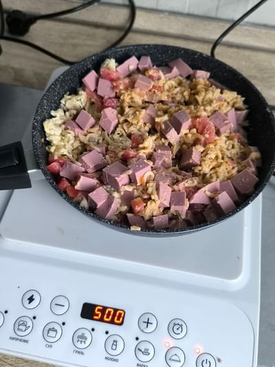 In the city of Ust-Luga, Russia, a frying pan filled with rice, diced sausage, and tomato is being cooked on a white electric induction cooker. The cooker displays a time setting of "500," likely representing minutes or a power level. Various cooking function icons are visible on the cooker's control panel, including those for boiling, soup, grilling, milk, and frying. The scene appears to be a domestic kitchen setting, with a power cord extending from the background.