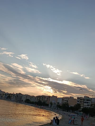 The image shows a beach scene at sunset in Águilas, Spain. The sky is a gradient of blue and orange, with sun rays breaking through the clouds. In the distance, buildings line the coast. In the foreground, people are scattered along the beach, some sitting, some walking, and some swimming. A bird is flying in the sky. A construction crane is also visible in the scene. The water is calm and reflects the orange light of the setting sun.