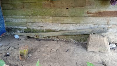 The image depicts a ground-level outdoor scene against a weathered wooden wall. A long, thick, weathered wooden beam rests horizontally on two supports in the lower middle section of the image. One support appears to be a roughly cut, circular wooden block covered with a white plastic bag, while the other is a large, trapezoidal stone block. The wooden beam has holes and appears worn. The ground is composed of packed dirt and debris. To the left, a blue tarp is partially visible, suggesting a sheltered area. A broken wooden post with green moss and a hole in it stands upright on the left side of the ground. A small, round stone is next to it. In the background, the wooden wall is constructed of horizontal planks, some with green algae or moss growing on them, indicating exposure to moisture. The overall scene suggests a rustic, possibly abandoned, outdoor space.
