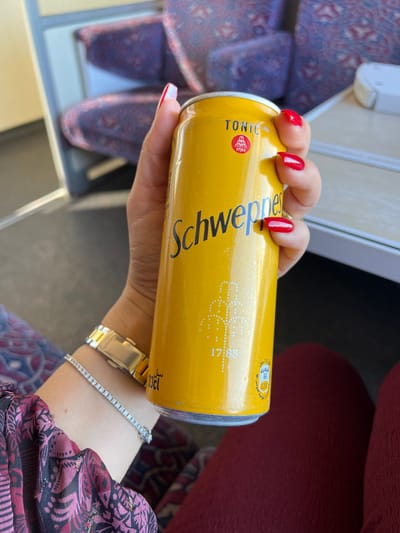 A close-up perspective captures a person's right hand holding a yellow Schweppes Tonic can inside a public transport vehicle in Sidi Kacem, Morocco. The hand displays bright red painted fingernails. On the wrist, a gold-toned watch with a visible clasp is worn alongside a thin, silver-colored bracelet featuring numerous small, reflective stones. The individual's clothing includes a patterned sleeve in dark red, pink, and black hues, and a segment of solid dark red fabric covering the lap. The Schweppes can is marked with 'Schweppes' in black script, 'TONIC' in capital letters, a red circular logo incorporating '1783', and a faint illustration of a fountain structure with '1788'. Additional details on the can include Arabic text and '33cl'. The background is out of focus, showing two upholstered seats with intricate blue, red, and beige patterns, characteristic of a train or bus interior. A light-colored interior structural element, likely a window frame or table edge, is partially visible to the right of the can.