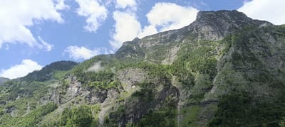 This is a wide-angle, low-angle shot of a steep, rocky mountain slope covered in dense green trees. The sky above is a vibrant blue with scattered white clouds. The mountain features rugged rock formations and varying textures, with some areas appearing smoother and others jagged. Patches of green vegetation cling to the rocky surfaces. In the distance, the mountain peaks rise dramatically against the sky. The overall scene depicts a natural, rugged mountain landscape, likely in the Austrian Alps, given the reference to Golling an der Salzach. There are no people or man-made objects visible in the image.