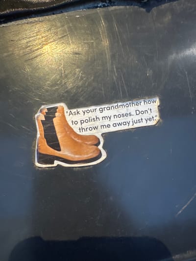 The media file displays a sticker on a dark, reflective surface that appears to be a piece of furniture or equipment. The sticker is predominantly white with a cut-out shape, featuring an illustration of two brown boots with black soles. Text is printed vertically on the white part of the sticker in black. The text reads: "Ask your grandmother how to polish my noses. Don't throw me away just yet." The lighting in the image suggests an indoor setting, with a strong light source creating reflections and highlighting scratches on the surface. There are no people visible, and the overall mood is neutral, conveyed by the static nature of the sticker and the mundane setting. The location context of Deventer, Netherlands, provides no specific visual cues within the image itself. The image is a close-up of the sticker and its immediate surroundings.