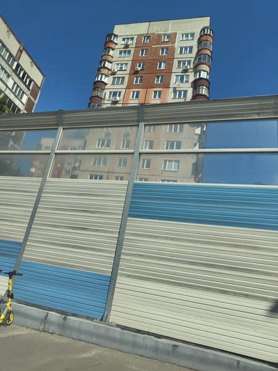 This is a medium-angle, eye-level, full exterior shot of a construction site or a sound barrier. The barrier consists of horizontal corrugated metal panels in shades of beige, light blue, and gray, with a section of transparent panels that reflect the sky and surrounding buildings. To the left, partially visible, is a yellow electric scooter. In the background, two apartment buildings are visible against a clear blue sky. The building on the right is taller and features a distinctive, curved section with circular balconies. The building on the left is simpler in design. The image appears to be taken in the city of Zhukovsky, Russia.