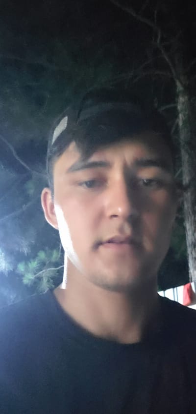 A close-up, vertically oriented, poorly lit, low-resolution selfie shows a young man from the chest up. He is wearing a black t-shirt and a black baseball cap with a white design on the front. His face is turned slightly towards the camera. He has dark hair, dark eyebrows, and is looking directly at the camera. His skin tone is light to medium. In the background, out of focus, are dark trees and some lights. The image appears to have been taken at night. The location is identified as Abdurahmoni Jomi, Tajikistan.