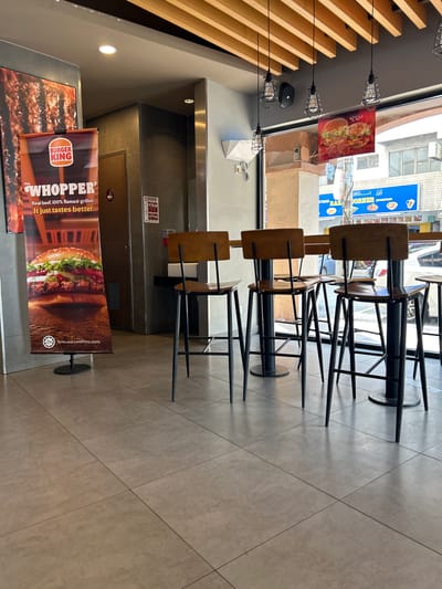 This image shows the interior of a Burger King restaurant located in Subang Jaya City Council, Malaysia. The scene is viewed from a low angle, looking towards a window and a counter area. In the foreground, there are several tall, dark-legged stools with wooden seats and backs arranged around a long wooden table. The floor is tiled in a neutral gray tone.
To the left, a large vertical banner advertises "THE WHOPPER" with an image of the burger and text stating "Real beef. 100% flamed-grilled. It just tastes better." A halal certification logo is visible at the bottom of the banner. Behind the banner, a door and a wall with tiled panels are visible.
In the background, through a large window, the exterior of the building and a street scene can be seen. A blue sign with Arabic writing and images of food items is mounted outside. The ceiling features exposed wooden beams and several pendant light fixtures. The overall environment suggests a modern, clean fast-food establishment. No people are visible in the image.