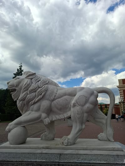 A marble statue of a lion with its paw resting on a sphere is prominently displayed outdoors. The lion is depicted in a powerful stance, with its mouth open and mane flowing. Behind the statue, a large building with red brick and decorative elements is visible, along with trees and a paved walkway. The sky above is filled with dramatic, grey clouds interspersed with patches of blue. The scene appears to be in a public space, likely a park or plaza, in Trafford, United Kingdom. There are a few indistinct figures in the background, suggesting a casual public gathering.