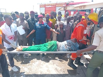 The image shows a scene in Katabu, Nigeria (latitude: 10.71, longitude: 7.514) where a man is being loaded into the back of a red fire truck. The man is lying on a stretcher, and two people are carrying him. There are a number of people standing around, some of whom appear to be onlookers and others who may be assisting with the loading process. The scene appears to be a street or parking lot, as there are vehicles and other people in the background. There is a woman in a blue uniform with a red cap who may be a first responder. The man on the stretcher is wearing a green shirt, green pants, and black sandals. The people carrying him are both wearing orange uniforms. The onlookers are dressed in various types of clothing, and most are wearing hats. The fire truck is partially open, with its rear door open. There is a large red "FEDERAL FIRE SERVICE" sign on the side of the truck. There is a fire hose lying on the ground in front of the truck. There is also a white sign behind the truck that reads "CHILE". The scene appears to be a medical emergency. It is not possible to identify the exact location in Katabu, Nigeria based on the coordinates provided.