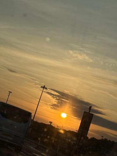 The image captures a scene in Uttlesford, United Kingdom. The sky is filled with clouds and a setting sun is visible. There are buildings, streetlights and trees on the right. A blue bus is parked in the foreground, partly obscuring the view. A road sign is visible above the setting sun.