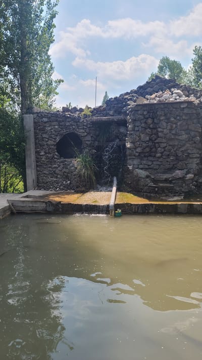 A stone structure with a small waterfall is depicted in Arstanbap, Kyrgyzstan. The structure appears partially ruined, with rough-hewn stones forming its walls and roof. A pipe protrudes from the waterfall, directing water into a murky pool in the foreground. Green foliage surrounds the structure, and a clear sky with scattered clouds is visible above. The scene appears to be an outdoor area, possibly a park or a natural attraction.