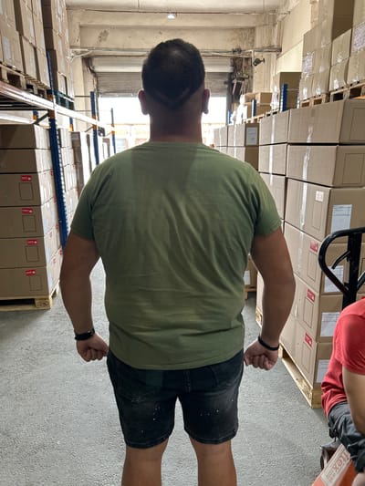 The media file shows a person in a warehouse, standing amidst stacks of cardboard boxes. The person is wearing a green t-shirt and dark shorts. The warehouse appears to be well-lit, with natural light coming from windows in the background. The boxes are neatly arranged on shelves and pallets, suggesting an organized storage environment. Some boxes have labels, but they are not legible enough to be read. A pallet with an "EPAL" logo is visible. The overall scene conveys a sense of industry and commerce, possibly related to logistics or retail. There are no clear indicators of time of day or weather conditions. The image is oriented vertically, with the person facing away from the camera.
