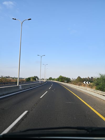 The image shows a view of a highway in Hod Hasharon, Israel, on a clear day. The road curves to the right, with a concrete barrier on the left and a metal guardrail on the right. Several lampposts are visible along the left side of the road, and directional arrows are placed on the right side. The road surface is asphalt with white lane markings and a yellow line marking the edge of the travel lane. The surrounding area is characterized by vegetation, including trees and bushes, and a distant hazy horizon. The view appears to be taken from inside a vehicle.