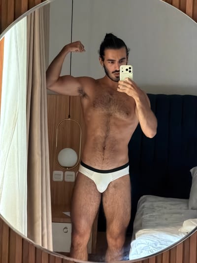 A man with dark hair pulled into a bun and a mustache is standing in front of a mirror in what appears to be a hotel room. He is wearing white briefs and has his left arm flexed. The room has a bed with white linens, a dark headboard, and a bedside lamp with a gold frame and a white globe. Curtains hang on the left side of the image. The image was taken in Pasir Putih, Indonesia.