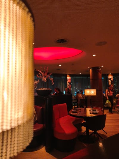 The image shows the interior of a dimly lit, upscale restaurant with a reddish-orange ambient glow. In the foreground on the left, a large, out-of-focus decorative light fixture with a woven texture dominates the left side of the frame. The middle ground features several tables with chairs, some occupied by patrons. A circular table with place settings is visible in the lower right quadrant. A woman in a pink top and dark pants is walking towards the right side of the image, carrying a green plastic bag. A large, round column with signage in Hebrew stands near the center. In the background, through large windows, a city skyline at night is visible, suggesting the restaurant is located in a high-rise building. The ceiling has a textured, dark finish with recessed lighting and a large, glowing red circular light fixture. The floor appears to be wooden. This scene is consistent with a restaurant located in a modern urban environment. While the image does not explicitly state it is in Be'er Sheva, Israel, the Hebrew signage on the column indicates a location where Hebrew is spoken, which includes Israel.