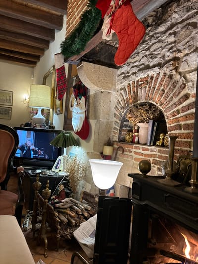 This media file captures a cozy, festively decorated rustic interior, likely a living room or common area, featuring stone and brick walls with exposed wooden beams. The scene is illuminated by artificial lighting, suggesting it is evening or night.

**Subjects/Objects:** A prominent fireplace dominates the right side of the image, constructed from rough-hewn stone and red brick, featuring a semi-circular brick arch. A fire burns brightly within the hearth. Above the fireplace opening, antique-looking brass decor is visible. Christmas stockings, one solid red and one plaid, hang near the fireplace, along with a green garland draped over a wooden beam. A Santa Claus figurine stands on a shelf below the fireplace, beside another plaid stocking. A stack of firewood is neatly piled next to the fireplace, topped with a newspaper.

In the center, a television screen displays a scene from a program, with partial French text visible: "...tout petit !". Various lamps provide illumination, including a tall floor lamp with a white bell-shaped shade near the fireplace and another lamp with a beige shade further left, next to a gold-framed mirror. Part of a dark armchair or sofa is visible in the foreground. A rustic wooden staircase with thick treads descends along the left side of the frame. An alcove in the brickwork above the fireplace contains dried foliage and small decorative items.

**Activity/Event:** The lit fireplace and Christmas decorations suggest a festive and warm atmosphere, likely during the holiday season. Someone may be relaxing and watching television.

**Notable Details:** The rustic architecture, including the stone and brickwork with wooden beams, implies an older, traditional building style, consistent with a rural or historic setting in Étréchy, France. The warm lighting and burning fire create a comfortable and inviting ambiance.

**Visible Text:** French text is visible on the television screen: "...tout petit !". Partially obscured French text, including "d'Anbass" and "questions ?", can be seen on the newspaper resting on the firewood.