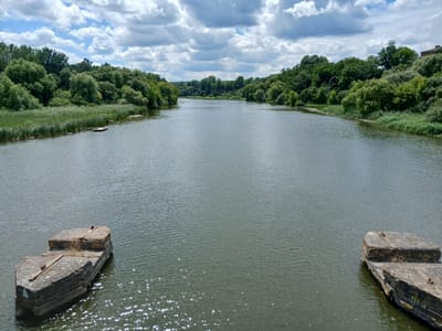 The image depicts a wide, calm river with green, tree-lined banks under a partly cloudy sky. In the foreground, two weathered concrete structures rise from the water on either side, suggesting remnants of a bridge or pier. The river flows towards a distant, hazy horizon, flanked by lush vegetation. On the left bank, tall reeds and smaller bushes are visible, and a small wooden raft or platform is partially submerged. The right bank features dense foliage and trees, with a hint of a building in the far distance. The water has a slightly choppy surface with reflections of sunlight. This scene is identifiable as the Irpin River in Stryzhavka, Ukraine, a location known for its natural beauty and recreational activities along the riverbanks. There are no people or animals visible in the frame.