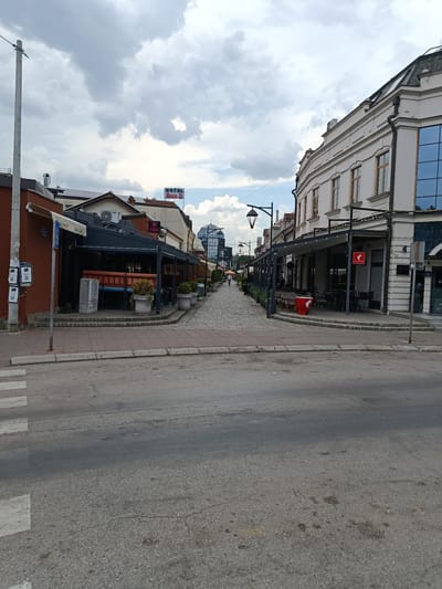 This is a wide-angle, eye-level shot of a cobblestone street in a European city, likely Niš, Serbia, given the architectural style and the presence of a "HOTEL DUO D" sign visible in the distance. The street is lined with buildings, many of which appear to be shops or restaurants with outdoor seating areas covered by awnings. The buildings vary in architectural style, with some older, ornate structures on the right and more modern ones on the left.
The scene depicts a quiet daytime, with no prominent activity. A few people are visible in the distance walking down the street, and some are seated at the outdoor tables of the cafes. The weather appears to be overcast, with a cloudy sky dominating the upper portion of the image. The clouds suggest a possibility of rain or recent rain, contributing to a muted light.
Notable details include street lamps lining the pedestrian walkway, potted plants placed outside the establishments, and a prominent red trash receptacle with a bird logo on it. A pedestrian crossing is visible in the foreground, indicating the street is also used by vehicles. The buildings on the right feature a classical architectural style with detailed facades and arched windows, while the buildings on the left are more contemporary, with some featuring modern glass elements. A distinctive spherical glass structure is visible further down the street, adding a modern contrast to the historic architecture. Several QR codes are displayed on a pole on the left side of the image. The overall mood is one of a calm, ordinary day in a city center.