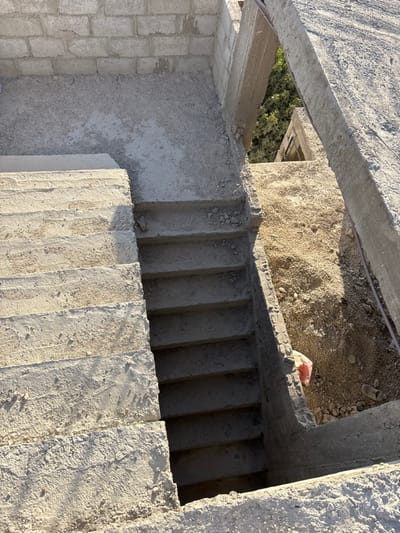 This image captures an unfinished construction site in Mu'adammiyat ash-Sham, Syria, under bright, direct sunlight. The scene is composed primarily of raw concrete and earth. In the left foreground, a flight of at least seven ascending concrete stairs, with a rough, untextured finish and visible aggregate, rises toward the upper left. Adjacent to these, a flat concrete landing area is covered with loose gravel and fine construction debris.
To the right of the landing, a flight of at least five descending concrete stairs, similarly untextured, leads into a shadowed opening. An excavated area of sandy-brown earth and loose gravel occupies the right side of the image, extending into the background. A partially obscured pink plastic bag is visible among the debris near the base of the descending stairs. Partial walls constructed from grey cinder blocks are visible in the upper left, while structural concrete beams and unfinished edges define other sections of the construction. Green foliage from trees or shrubs is visible through an opening in the construction in the upper right background, suggesting a surrounding outdoor environment. No individuals, actions, or interactions are present in the scene.