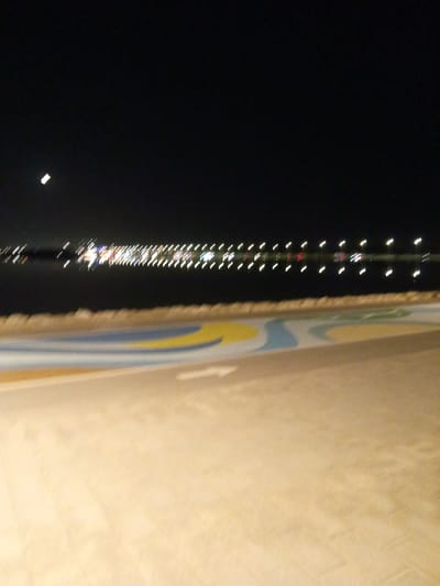 The image shows a nighttime view of a waterfront promenade in Al Khobar, Saudi Arabia. The foreground features a paved pathway with a colorful, abstract design of yellow, blue, and white curved lines. Beyond the pathway, there is a rocky embankment, and then a dark body of water. Across the water, a long line of lights, likely from buildings or a bridge, stretches into the distance, reflecting on the water's surface. In the upper left corner of the black sky, a faint crescent moon is visible. The image appears to be taken at a slight distance from the water's edge.