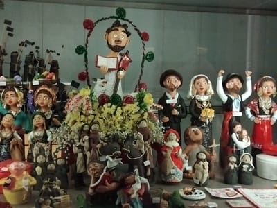 The image depicts a collection of religious figurines displayed in a shop window in Braga, Portugal. The figurines, predominantly made of clay or ceramic, represent various biblical figures and saints. In the center, a larger figure of a man holding a book and a cross is prominent, surrounded by smaller figures of animals, including a sheep. To the right of this central figure, a couple dressed in traditional attire, possibly representing a wedding or a celebratory event, stands with raised hands. Further to the right, another female figure in a red dress is visible, holding what appears to be a bundle of wheat. The background shows more figurines in various poses, some of which appear to be engaged in religious activities or daily life. The display is decorated with artificial flowers and greenery, enhancing the festive and devotional atmosphere. The overall arrangement suggests a display of folk art or religious imagery typically found in religious shops or markets in Braga, a city known for its rich religious heritage.