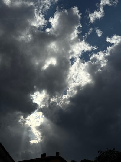 An eye-level, upward-angled shot reveals a dynamic sky. To the left, patches of bright blue sky are punctuated by small, white, puffy clouds. Dominating the center and right of the frame are large, ominous storm clouds, ranging in shade from gray to nearly black. The sun peeks through a gap in the clouds near the center-right, casting radiant sunbeams. Parts of a building and tree foliage are in the bottom-right corner of the image. The photograph was taken in Zhaltusha, Bulgaria.