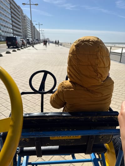An overhead, slightly tilted shot captures a person, likely a child, seated in a pedal go-kart on a sunny day. The individual is seen from behind, wearing a hooded, mustard-yellow quilted jacket. Their hands are not visible on the black steering wheel of the go-kart, which features a blue and yellow metal frame and a black seat. The go-kart appears well-used, with some paint wear on its black and blue components.
The setting is an outdoor promenade or boardwalk, indicated by the paved ground and the presence of buildings to the left and a distant horizon line to the right, suggesting the sea. The sky is bright and clear blue, indicating fair weather during daytime. In the background, tall, modern buildings line the promenade, and street lamps are visible. Several blurry figures of people can be seen walking further down the promenade.
A notable detail is a black panel on the go-kart's body, which displays text: "ZEEDIJK 122 - MIDDELKERKE - TEL 059 30 62 93 - GSM 0476 36 39 25". This text confirms the location as Middelkerke, Belgium, specifically at number 122 on the "Zeedijk" (sea dike or promenade), and provides contact information. The image conveys a leisurely outing by the sea.