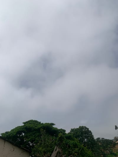 The image captures a view from a residential area in Zhwarki Mile 10, Nigeria, under an overcast sky. The foreground features the tops of lush green trees and the upper portion of a building with a textured, possibly concrete, wall. The sky is predominantly filled with grey clouds, with some patches of lighter sky visible. In the distance, silhouetted against the sky, are more trees and a hint of a landscape suggesting rolling hills. The scene appears to be quiet and undeveloped, typical of a suburban or semi-rural setting within the city. There are no people or specific actions depicted.