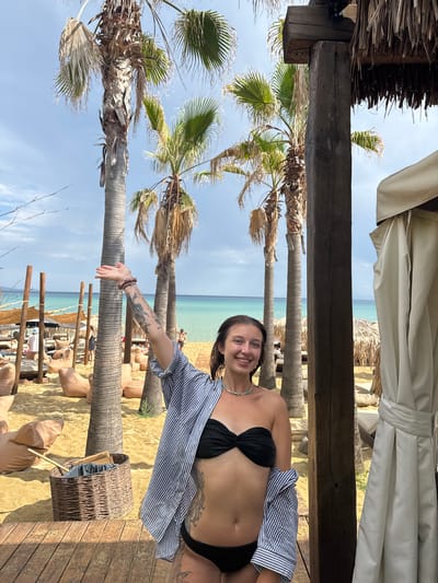 A young woman with a tattoo on her arm is standing on a wooden deck, smiling at the camera. She is wearing a black bikini top and a blue and white striped shirt. She is standing in front of a beach with palm trees and beanbag chairs. The ocean is visible in the background. The location is Nea Peramos, Greece.