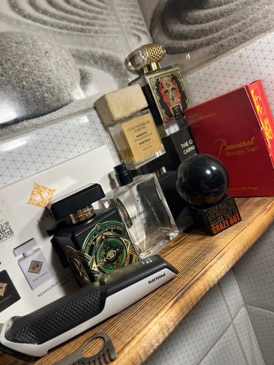 The image shows a collection of perfume bottles and a red box, possibly related to fragrances, placed in a wooden drawer. The items are arranged on a patterned surface. Visible text includes "Baccarat Rouge 540," "CRAZY HOT," "oldschoolkiller," "THE GREEN CARNATION," "MUSK THERAPY," and "PHILIPS." The overall impression is one of personal grooming or cosmetic products. The lighting suggests an indoor setting, and the time of day is not discernible. The location context provided is "Balaklavsky District, Ukraine," which may or may not be directly represented by the visual content. The scene appears to be a still life arrangement of these items.