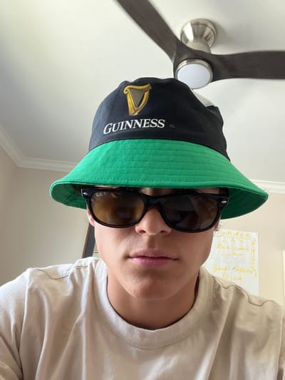 The image presents a close-up, chest-up view of a young individual, positioned indoors in Rancho Palos Verdes, United States. The subject, of indeterminate gender, wears a black and green bucket hat prominently displaying a golden harp emblem and "GUINNESS" text. Dark, polarized Ray-Ban Wayfarer-style sunglasses, with "Ray-Ban P" visible on the left lens, obscure the subject's eyes. A plain, light-colored (beige or tan) short-sleeved t-shirt covers the torso. The subject's mouth is closed, and the facial expression is neutral, directed towards the camera.
The background elements include a white ceiling with crown molding in the upper left. A three-bladed ceiling fan with a brushed metal housing and a central white light fixture is visible in the upper right portion of the frame. To the right of the subject, a white board displays handwritten text in a light-colored marker, partially legible, including "SUMMER", "LAX", "Rock-climbing", "Guitar", "Hybrid Athlete", and "YOGA". A dark-framed object, possibly a picture or mirror, is partially visible behind the subject's left shoulder. The scene is brightly illuminated.