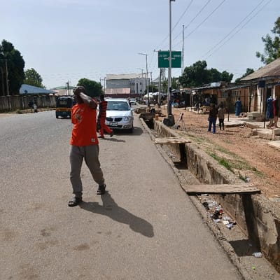 The scene is a street in Kufang, Nigeria. A man in an orange shirt and gray pants is walking away from the camera, holding his right hand over his head. He is walking on a paved road with a drainage ditch running parallel to it on the right. On the right side of the road, there are several buildings with open storefronts. In the foreground, a man with a black shirt and red stripe is walking away from the camera. There is a white car parked on the right side of the road, facing the camera. A green street sign with "JOC CITY" and an arrow is visible on a pole above the ditch. There are also several people walking in the distance. The sky is clear and blue, with a few white clouds. The ground is dirt with a few trees and shrubs in the distance. The scene is an urban area in Nigeria, with evidence of both modern and traditional infrastructure.