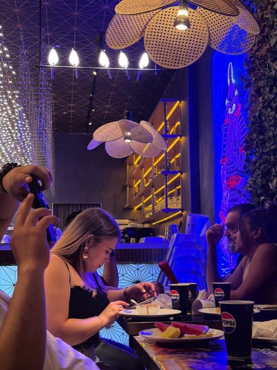 The image depicts a group of people at an indoor establishment, likely a restaurant or bar, as indicated by the presence of tables, drinks, and food. The setting appears modern and stylish, with a prominent neon light display of a dragon on the ceiling, illuminated in vibrant blue. Decorative woven light fixtures hang from above, casting a warm glow. In the foreground, a woman with blonde hair is visible, wearing a black top. Her hands are engaged with a black device, possibly a phone or remote.
Several Pepsi cups are arranged on a table, along with plates of fruit, including watermelon and pineapple. The presence of these items suggests a dining or social gathering. The overall lighting is dim, with the neon lights and overhead fixtures providing illumination, creating a moody ambiance. The time of day is not explicitly indicated, but the indoor setting and lighting suggest it could be evening or night.
The image is oriented vertically, and the people and objects are seen from a slightly elevated perspective. The people's faces are partially visible, but their emotions are not clearly discernible. There is text visible on the Pepsi cups, reading "PEPSI". The scene evokes a sense of casual social interaction within a contemporary, well-lit environment.