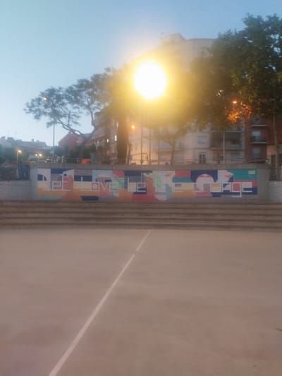 The image depicts a public space in Rubí, Spain, possibly a park or recreation area. A concrete surface, possibly a basketball court or plaza, occupies the foreground, with a white line visible. A set of steps leads up to a wall adorned with a colorful mural with the words "LOVE PEACE". Behind the wall, trees and buildings are visible, silhouetted against the bright sky. A light source in the upper center, possibly the sun, creates a strong glare, obscuring some details. The scene appears to be captured around dusk or dawn due to the subdued lighting. No people are directly visible in the image.