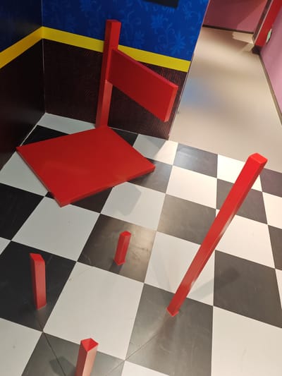 The image depicts an optical illusion room with a checkered floor made of black and white squares. Several red geometric objects are placed on the floor. In the foreground, there are three short, rectangular pillars and a long, thin rectangular beam. In the midground, a large, flat red square is positioned on the checkered floor. In the background, a red structure resembling a flag pole with a rectangular flag is attached to a wall. The walls are decorated with a blue patterned wallpaper and a yellow stripe. The overall scene creates a sense of distorted perspective, typical of an Ames room, where objects appear to change size as they are moved. There is no indication of people, specific time of day, weather, or emotions. The location context provided is Gongshu District, China. No visible text is present in the image.