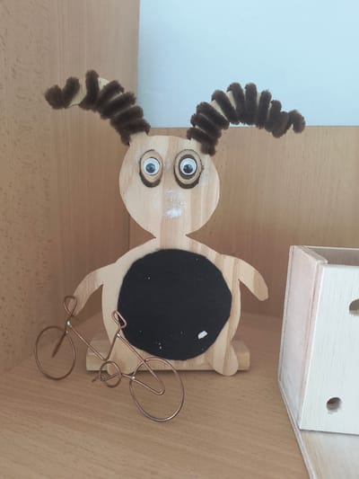 This media file shows a close-up, top-down view of a wooden craft item resembling a cow. The cow is made from light-colored wood and has two googly eyes, one white and one black. It has brown pipe cleaner horns that curl outwards and downwards, and a black felt circle on its body, possibly representing a patch or design. There is also a small white speck on the wood near the eyes. Attached to the side of the cow is a small, decorative copper wire bicycle. The craft is positioned on a light brown wooden surface, which appears to be a shelf or tabletop. In the background, there is a light-colored wooden structure with visible circular holes, possibly part of a larger toy or display. The lighting suggests an indoor setting, possibly during daytime. The image is cropped, showing only the craft item and its immediate surroundings. There is no visible text in the image. The overall scene is a still life of a handmade object.
