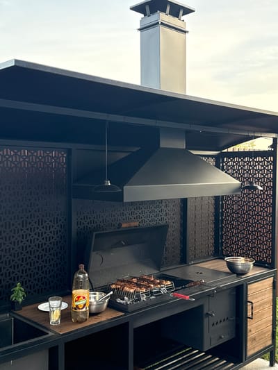 This image captures an outdoor barbecue setup in what appears to be a backyard or patio area in Starokorsunskaya, Russia. The main subject is a large, modern black metal barbecue grill structure. It features a prominent black range hood, a grilling area with a rack of skewered food, and various shelves and compartments. To the left, a tall, silver metal chimney or ventilation pipe extends upwards. On a wooden shelf to the right, there are several items: a metal bowl, a large plastic bottle of oil with a yellow and red label, a small shot glass on a saucer, and a small potted plant. Above the grill area, a decorative metal screen with a geometric pattern adds visual interest. The overall scene suggests a preparation for or the active process of grilling, indicated by the food on the skewers. The lighting suggests it is daytime, possibly late afternoon, with a bright, overcast sky visible in the background. The setting is a residential outdoor space, with wooden elements and metal construction of the barbecue unit being the most prominent features. There is no visible text other than what is on the oil bottle label, which is not clearly legible.