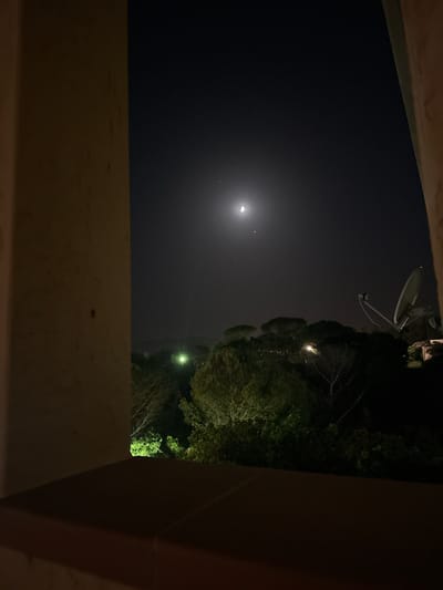 The image displays a night scene, possibly viewed from a window, looking out over a landscape. In the sky, a crescent moon is visible, accompanied by two smaller celestial bodies. Below the sky, a dark silhouette of trees and foliage dominates the middle ground. Scattered among the trees are faint, artificial lights, one of which appears bright green. On the right side of the frame, a satellite dish stands out against the dark sky and trees. The foreground is partially obscured by a warm-toned, textured surface, likely a windowsill or ledge, and a portion of a wall or pillar on the left. The overall impression is of a rural or semi-rural environment at night, potentially in or near Biddeputzi/Villaputzu, Italy, given the context. No people or specific actions are depicted.