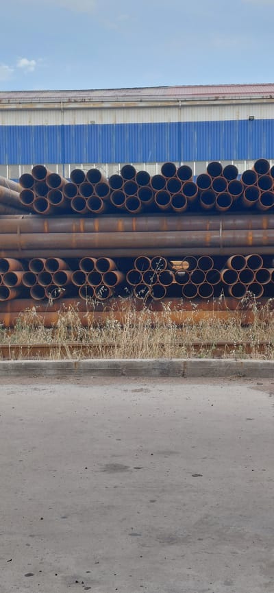 In Ohangaron, Uzbekistan, a stack of industrial pipes is stored outdoors. The pipes are made of metal and appear to be rusty. They are arranged in multiple horizontal layers, with some layers forming compact bundles and others more spread out. In the background, a building with a blue corrugated metal facade and a white and red roof is visible. The sky above is partly cloudy. The ground in the foreground is an asphalt surface.