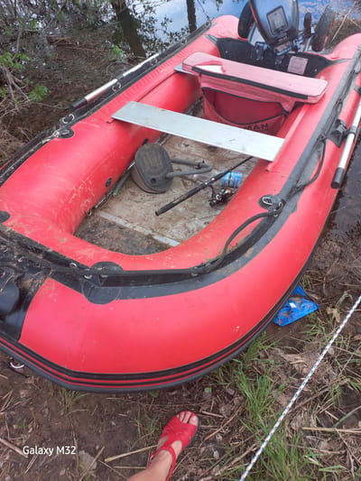 A red inflatable boat rests partially in the water and partially on the shore. The boat is equipped with a small outboard motor and oars. Various pieces of equipment, including fishing rods, are inside the boat. A person wearing a red sandal is visible near the edge of the boat. The surrounding environment consists of grass, mud, and trees. The photo was taken on the banks of the Akhtuba River in the city of Volzhsky, Russia.