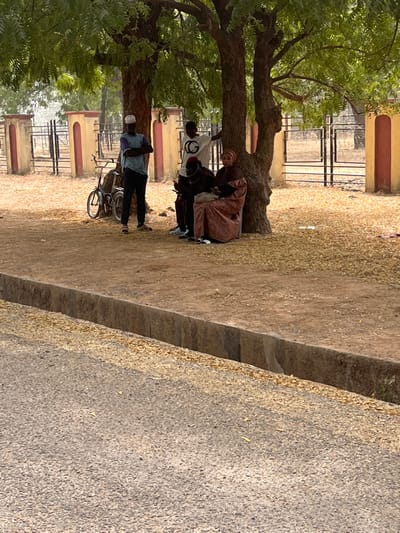 The image depicts a roadside scene in Kanti, Nigeria. Five individuals are gathered under the shade of two trees on a dirt patch adjacent to a paved road.
From left to right:
* A male figure stands with arms crossed, wearing a light blue long-sleeved shirt, dark trousers, white sandals, and a white skullcap. A dark bicycle is parked behind him, leaning against the tree trunk.
* Behind him, partially obscured by the tree, another male figure in a white t-shirt with a "CG" logo and dark trousers stands.
* Next to the tree, a male figure is seated on the ground, wearing a dark shirt, dark trousers, and a red and black cap. He appears to be looking down at a mobile device held in his hands.
* To his right, a female figure is seated, wearing a patterned reddish-brown long dress (hijab) and a light brown head covering. She holds a small dark object in her lap.
* A fifth person, a child, is visible between the seated male and female, partially obscured.
The ground under the trees is dirt, covered with numerous dried leaves. Behind the individuals, there is a low concrete and metal fence with light-colored pillars. In the foreground, a paved road is visible, separated from the dirt patch by a concrete curb and a narrow, open ditch. The lighting suggests a sunny day, with dappled shade cast by the trees.
