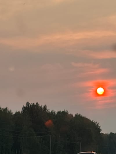 An image taken in Staryi Poros, Russia, depicts a sunset with the sun appearing as a bright orange orb surrounded by a reddish glow. The sky is filled with hazy clouds in shades of orange and pink. In the foreground, a dark line of trees forms a silhouette against the sky. Power lines and poles are visible in front of the trees. The bottom of the image shows the roof of a white vehicle.