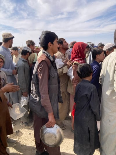 This outdoor image, captured in Chaman City Tehsil, Pakistan, depicts a dense gathering of approximately 15-20 individuals, primarily adult males and adolescent boys, illuminated by natural daylight under a partially cloudy sky. The ground surface is unpaved dirt.
In the foreground, a young male, appearing adolescent, stands center-left, wearing a dark brown long-sleeved shirt and a dark grey sleeveless vest. He holds an empty, polished metallic bowl in his right hand. To his immediate right, a young boy is visible from the back, wearing a long, dark-colored tunic. Directly behind the foreground male, another man in a light brown striped shalwar kameez and a light-colored scarf holds a small white plastic bag. Further right, a man with a red head covering is visible in the mid-ground.
To the left of the foreground male, an adult male wears a light blue shalwar kameez and a beige kufi cap, holding a clear rectangular plastic container in his left hand. Other individuals in the mid-ground and background are dressed in various traditional attire, including shalwar kameez and kufi caps, with several also holding small white plastic bags or clear plastic containers. Many individuals are looking generally towards the right side of the frame or upwards, suggesting attention towards an unseen area.
The background reveals utility lines or wires against the sky, with indistinct structures or geographical features in the far distance. Shadows suggest a bright, daytime setting.
