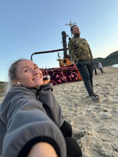 The image depicts a beach scene in Teriberka, Russia, featuring a rusted shipwreck as a prominent element. In the foreground, a woman with blonde hair pulled back is smiling and looking at the camera, holding a white object to her cheek. She is wearing a gray fleece jacket and white wireless earbuds. Behind her, a man with a beard, wearing a camouflage jacket and dark pants, stands with his hands in his pockets, looking upwards. In the background, more people are visible walking on the sandy beach, with hills and the ocean in the distance. The sky is clear and blue, suggesting daylight. The shipwreck, covered in graffiti, is partially submerged in the sand.
