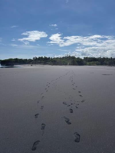 The image shows a black sand beach in Selemadeg Timur, Indonesia. The beach stretches horizontally across the frame, with a line of trees separating it from a blue sky with scattered white clouds. Footprints are visible in the sand, leading away from the viewer. The beach appears deserted except for the footprints.